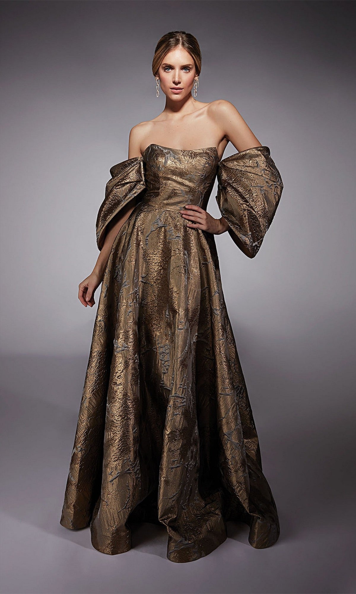 A woman models the Alyce 27760 long formal dress in bronze, featuring a strapless neckline and voluminous off-the-shoulder sleeves, set against a plain gray background.